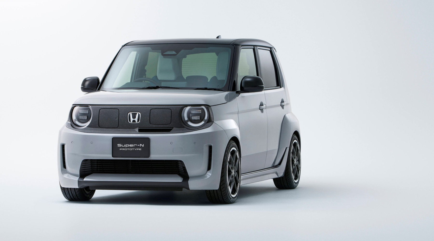A Honda Super-N prototype, a compact electric vehicle with a boxy design. The car is light gray with a black roof, featuring round LED headlights and a minimalist front grille with the Honda logo. It has a modern, urban-friendly appearance with clean lines and sporty alloy wheels.